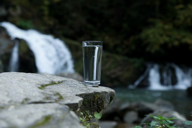 Photo of Glass of fresh water on stone near waterfall outdoors