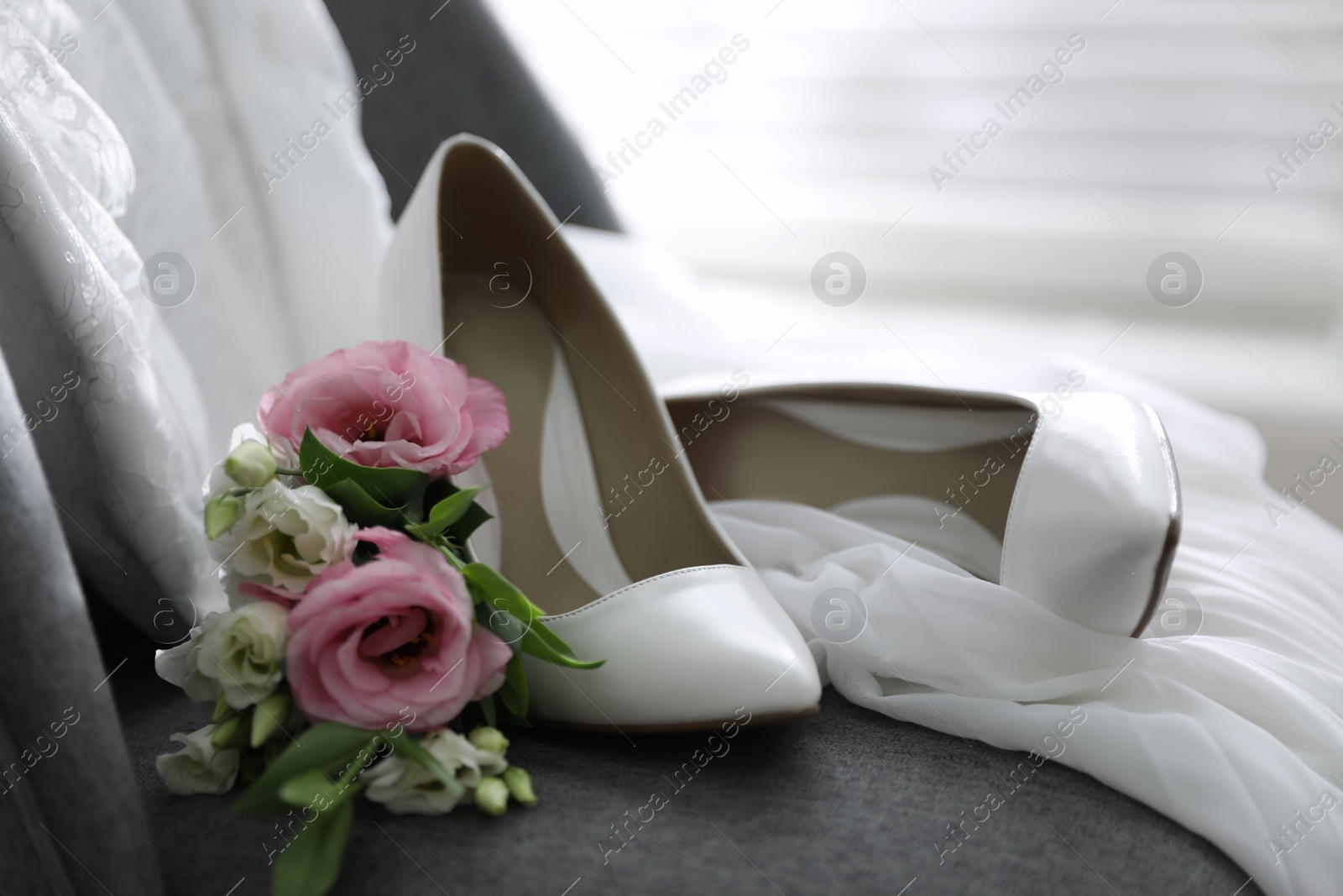 Photo of Pair of white high heel shoes, flowers and wedding dress on chair indoors