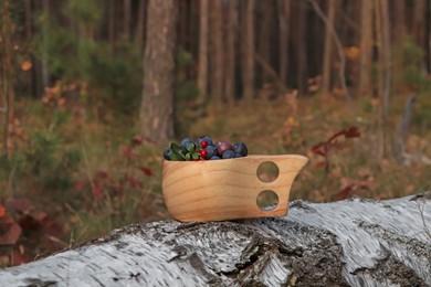 Photo of Wooden mug full of fresh ripe blueberries and lingonberries on log in forest