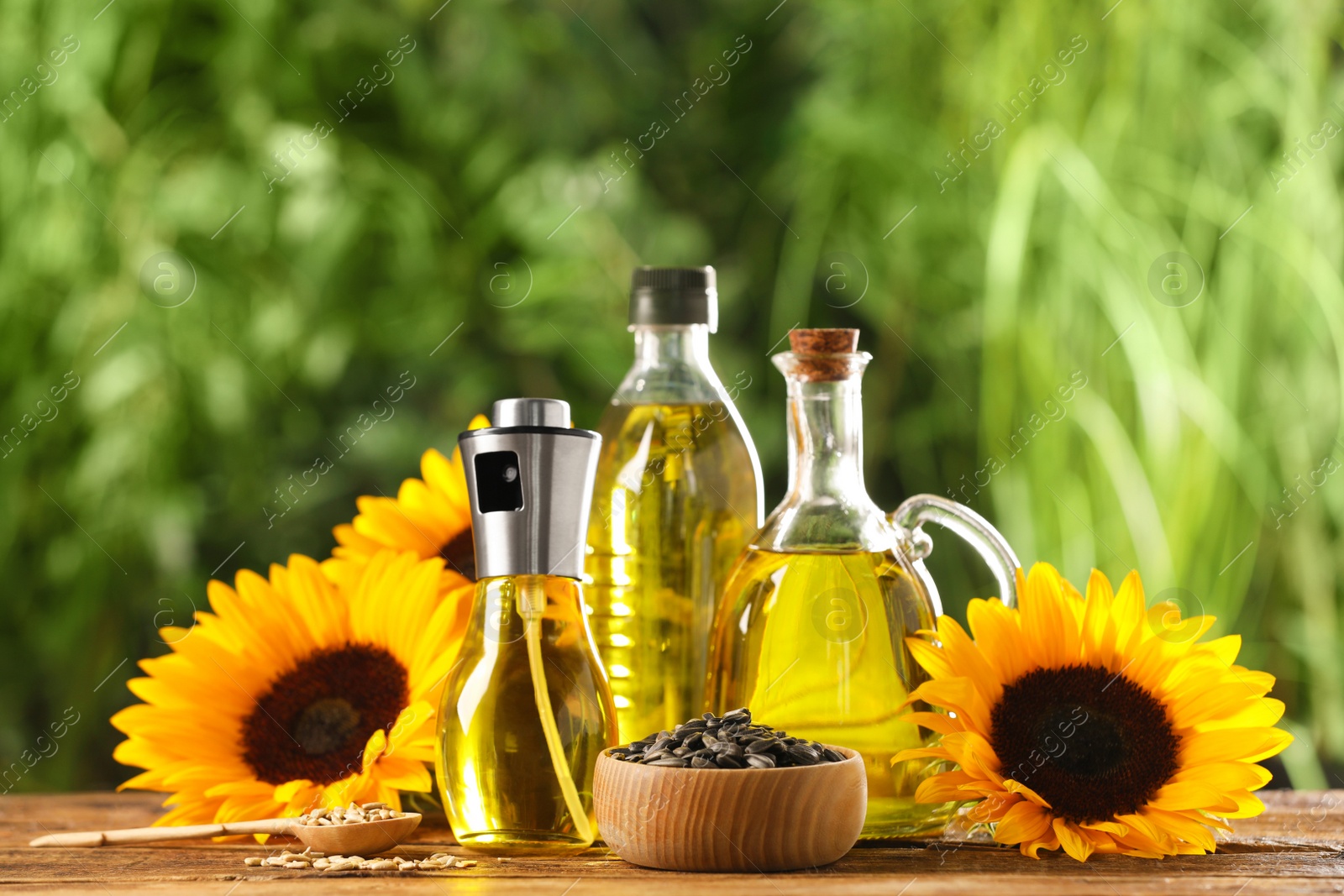 Photo of Many different bottles with cooking oil, sunflower seeds and flowers on wooden table against blurred background