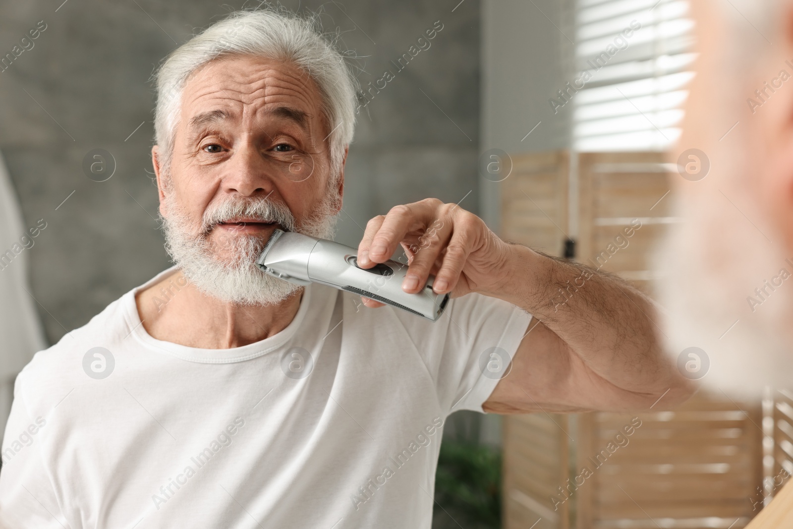 Photo of Senior man trimming beard near mirror in bathroom