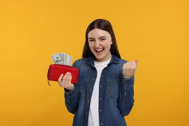 Excited woman holding wallet with dollar banknotes on orange background