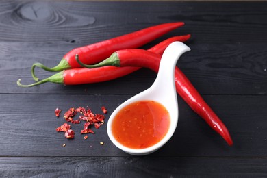 Spicy chili sauce in spoon and peppers on black wooden table, closeup