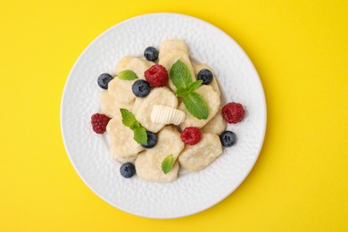 Plate of tasty lazy dumplings with berries, butter and mint leaves on yellow background, top view