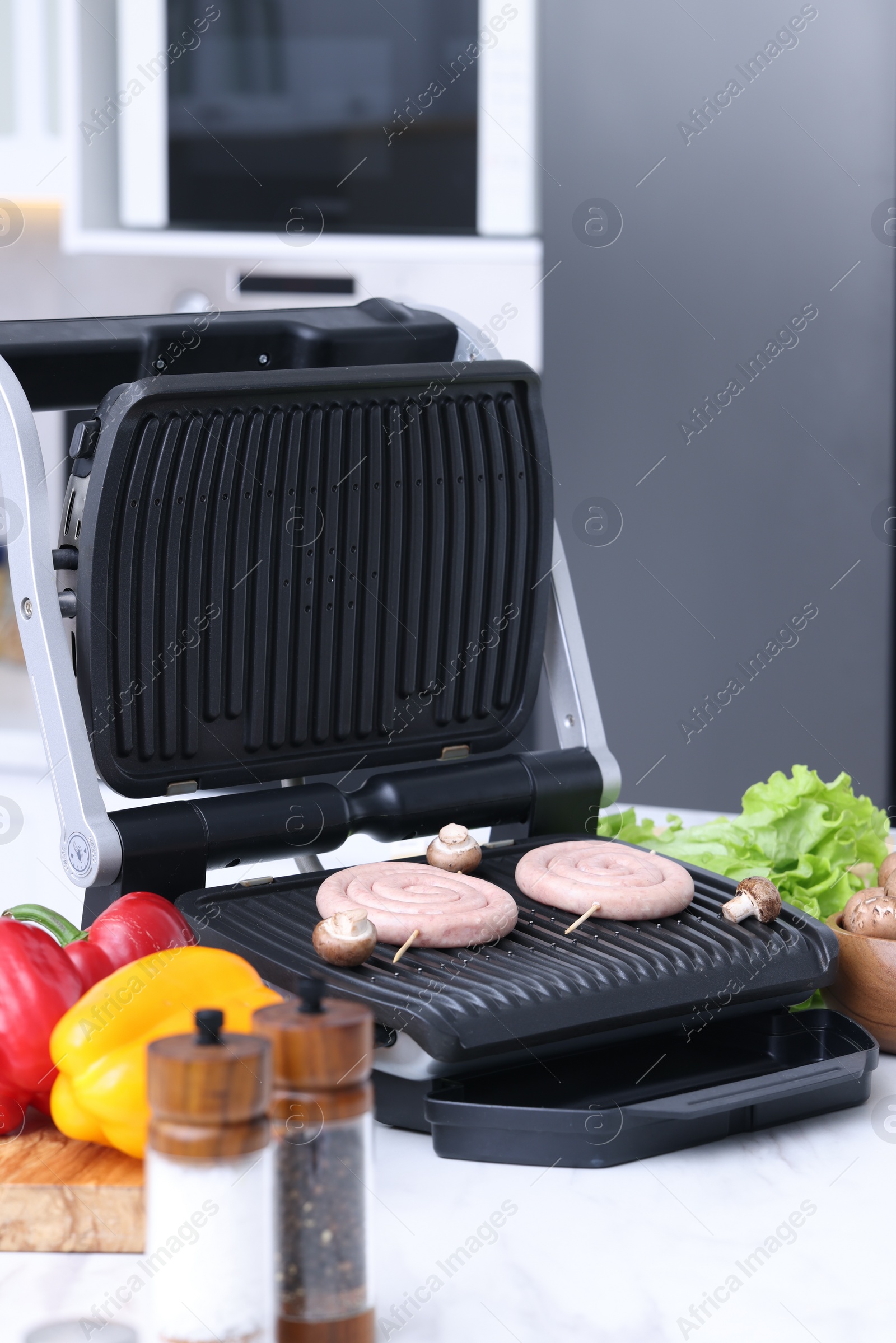 Photo of Electric grill with homemade sausages, mushrooms, vegetables and spices on white table in kitchen