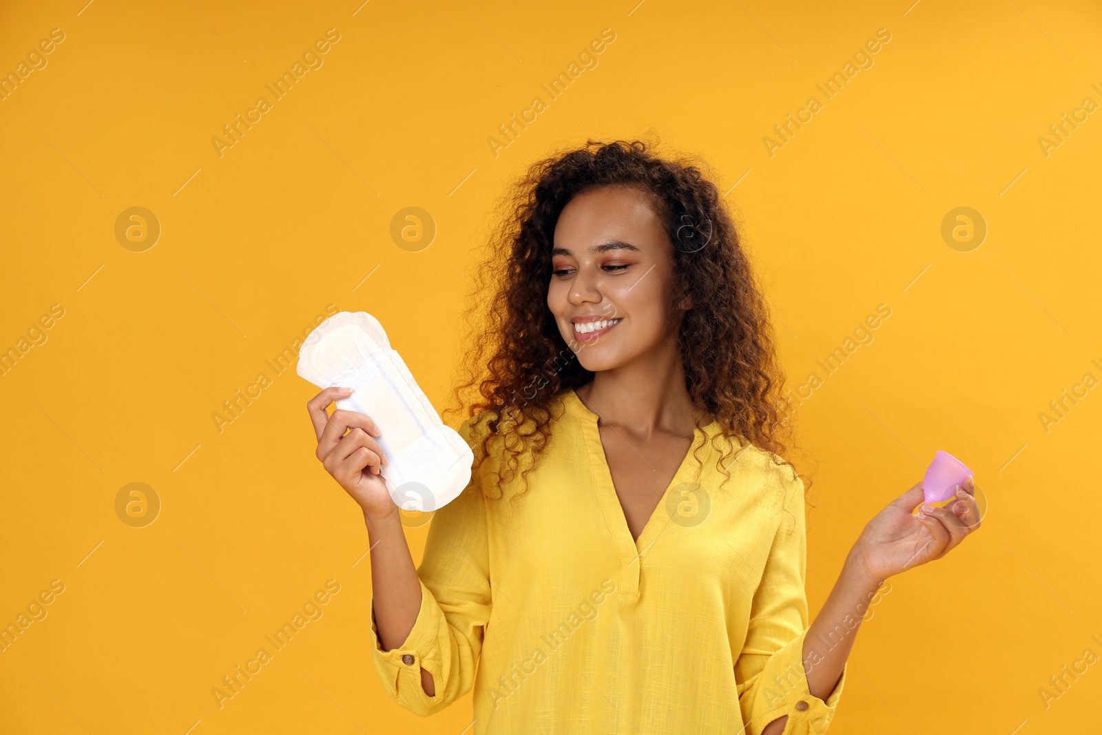 Photo of Young African American woman with menstrual cup and pad on yellow background