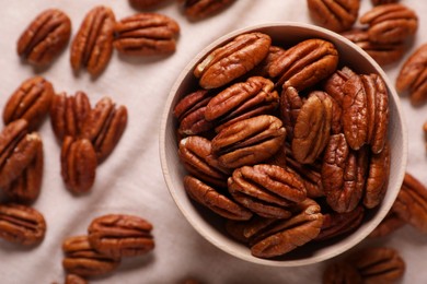 Tasty pecan nuts with bowl on beige cloth, flat lay