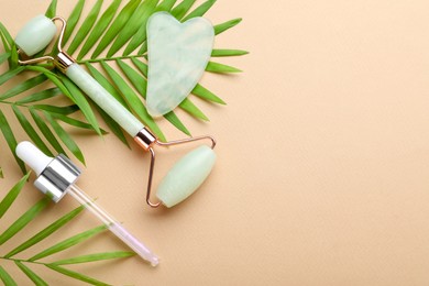 Gua sha stone, face roller, dropper and green leaves on beige background, flat lay. Space for text