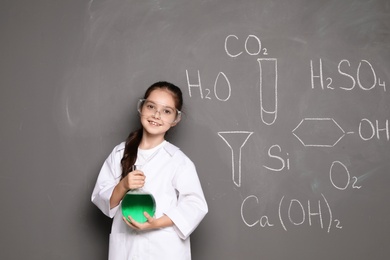 Little school child in laboratory uniform with flask of liquid and chemical formulas on grey background