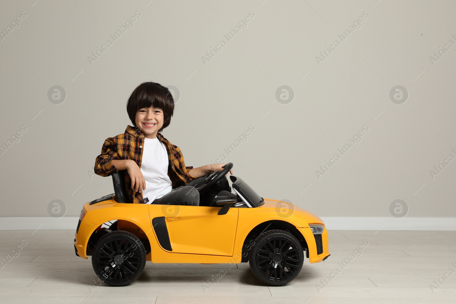 Photo of Cute little boy driving children's electric toy car near grey wall indoors. Space for text