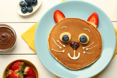 Photo of Creative serving for kids. Plate with cute cat made of pancakes, berries, cream, banana and chocolate paste on white wooden table, flat lay