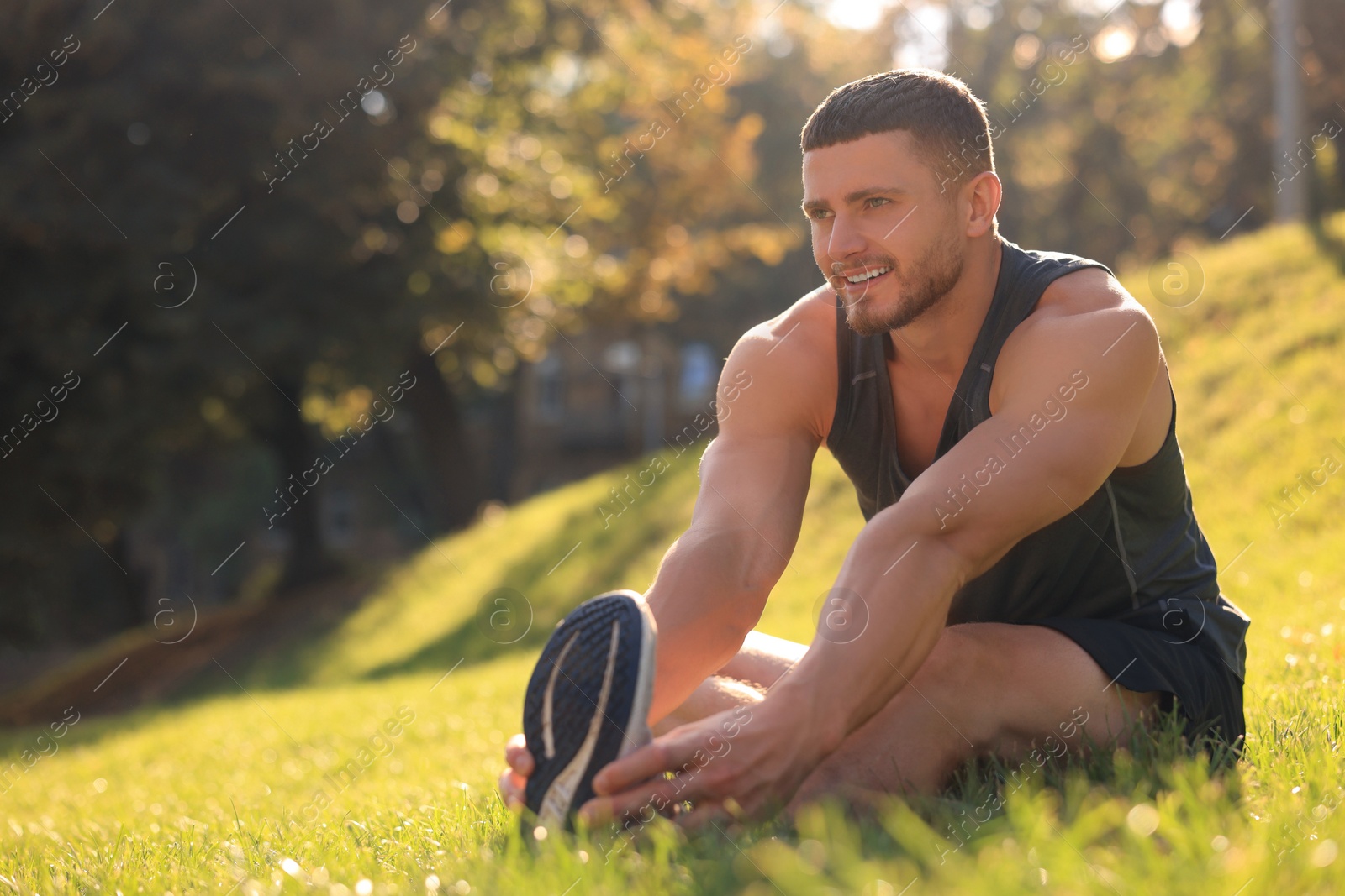 Photo of Attractive man doing exercises on green grass in park, space for text. Stretching outdoors