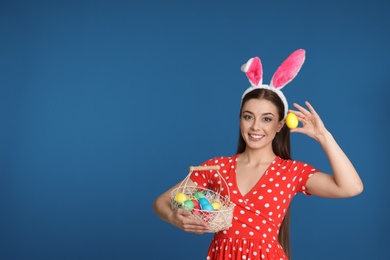 Beautiful woman in bunny ears headband with Easter eggs on color background, space for text