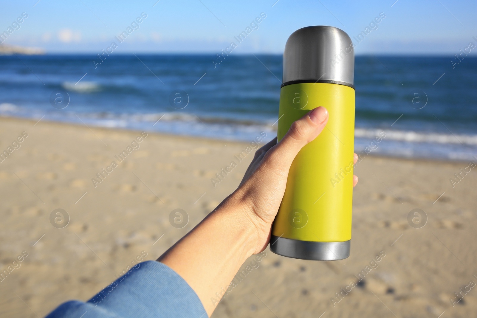 Photo of Woman holding yellow thermos with hot drink on beach near sea, closeup. Space for text