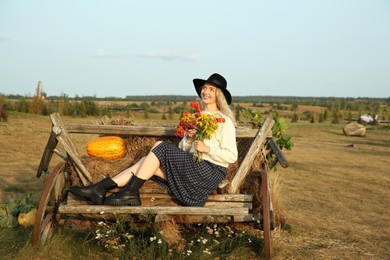 Photo of Beautiful woman with bouquet sitting on wooden cart with pumpkin and hay in field. Autumn season