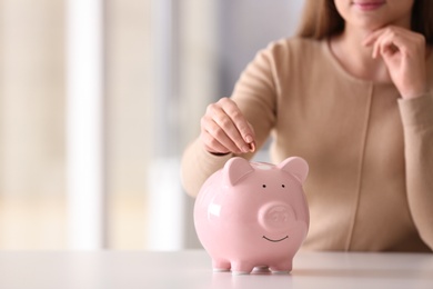 Woman putting coin into piggy bank at table indoors, closeup. Space for text