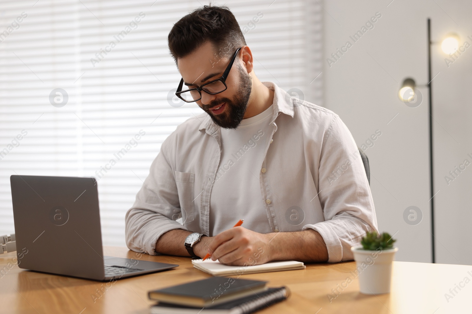 Photo of Young man writing down notes during webinar at table in room