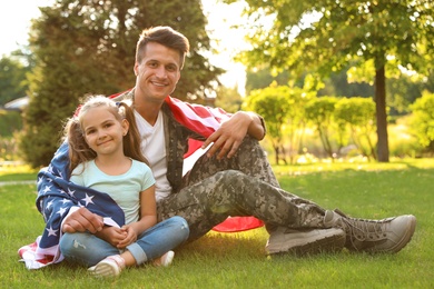 Father in military uniform with American flag and his little daughter sitting on grass at park