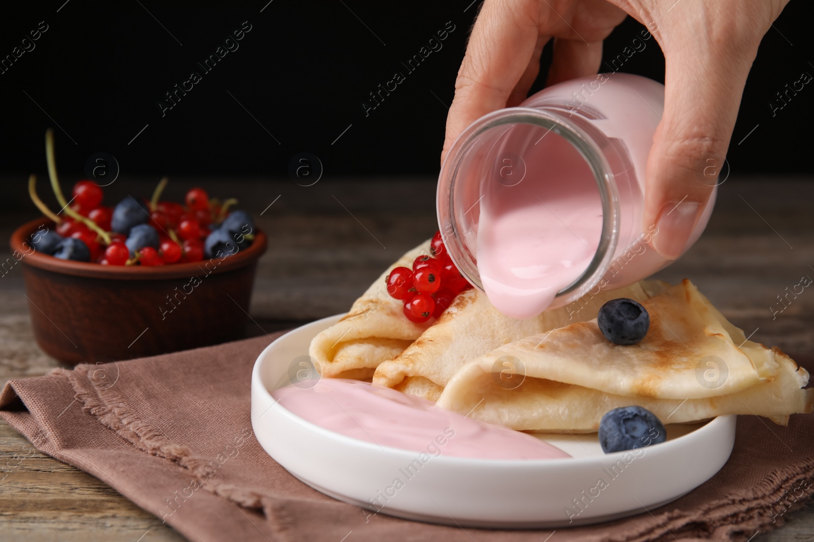 Photo of Woman pouring natural yogurt onto crepes with blueberries and red currants at wooden table, closeup