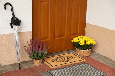 Photo of Door mat with word Welcome, umbrella and beautiful flowers near entrance