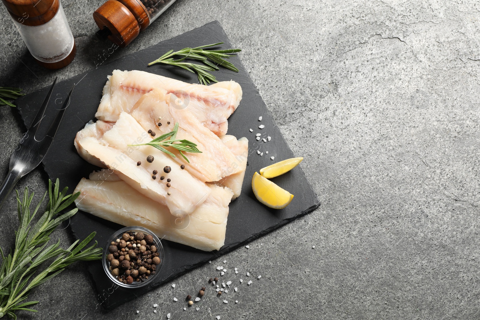 Photo of Pieces of raw cod fish, rosemary, peppercorns and lemon on grey table, top view. Space for text