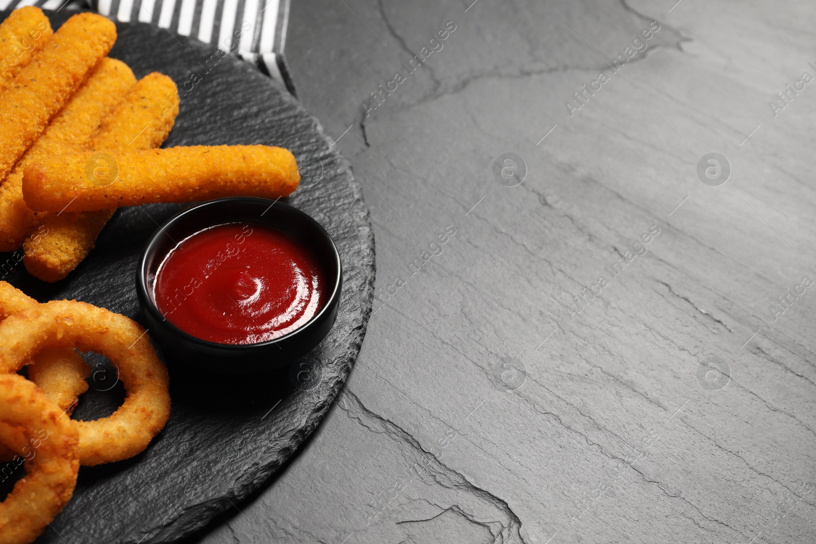 Photo of Tasty ketchup, onion rings and cheese sticks on grey textured table, closeup. Space for text