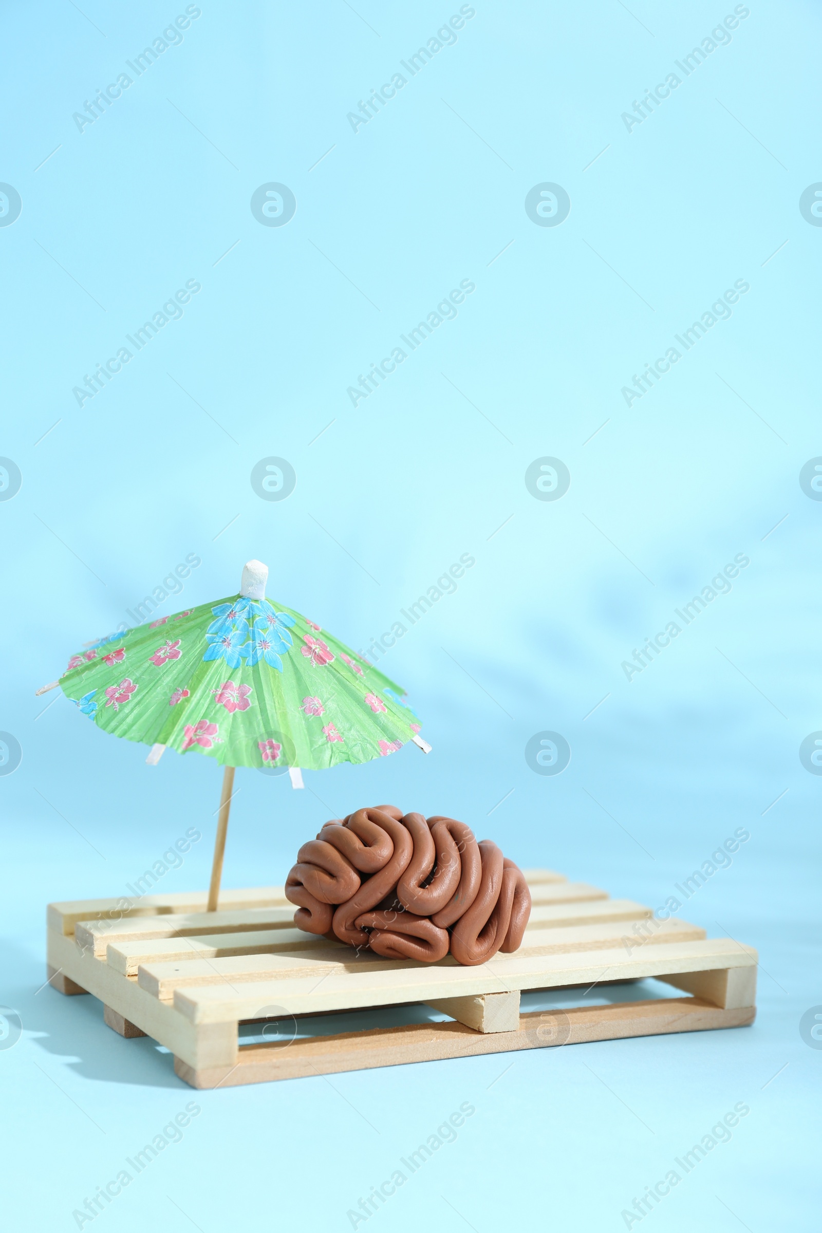 Photo of Brain made of plasticine on mini wooden sunbed under umbrella against light blue background, space for text