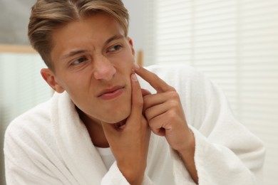 Photo of Upset young man looking at mirror and popping pimple on his face indoors. Acne problem
