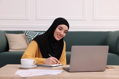 Photo of Muslim woman in hijab writing notes near laptop at wooden table indoors