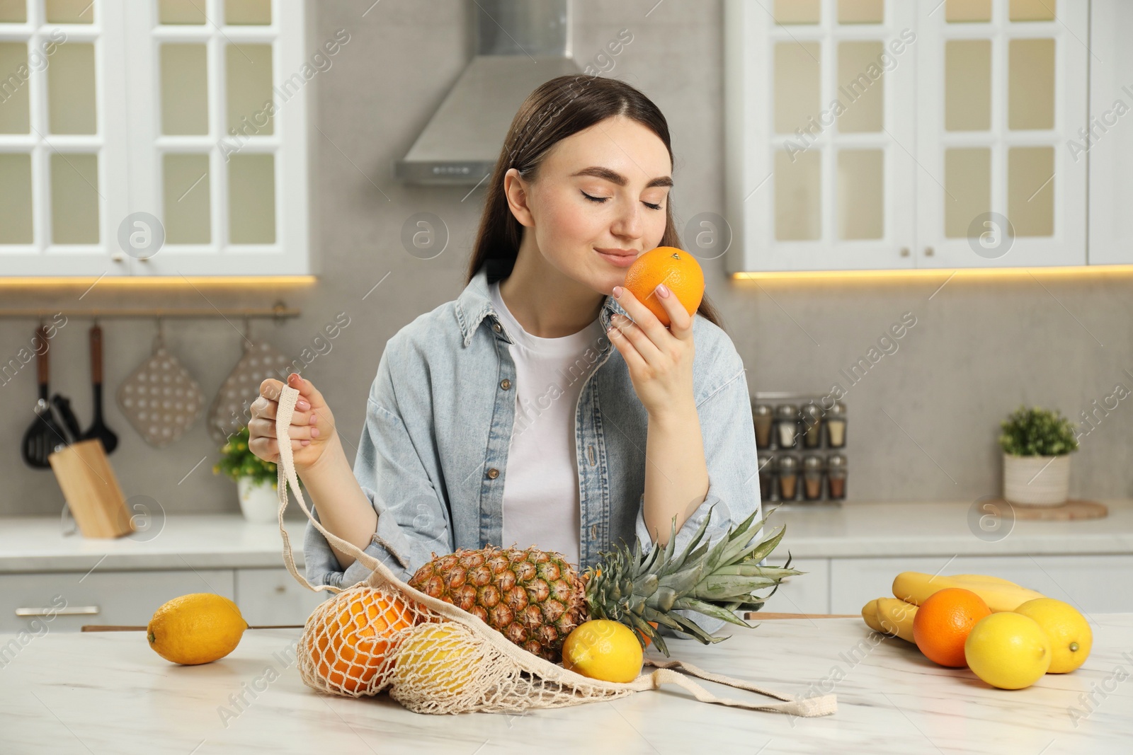 Photo of Woman with string bag of fresh fruits at light marble table in kitchen