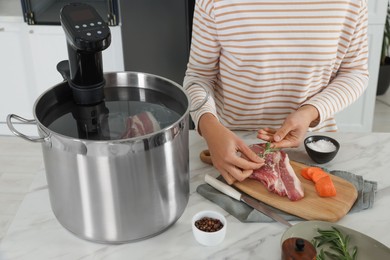Photo of Woman adding rosemary to meat near pot with sous vide cooker at table, closeup. Thermal immersion circulator