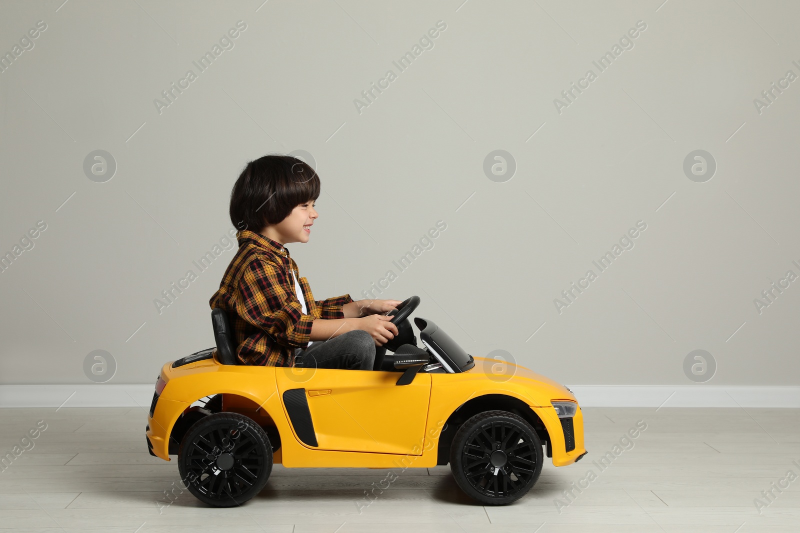 Photo of Cute little boy driving children's electric toy car near grey wall indoors