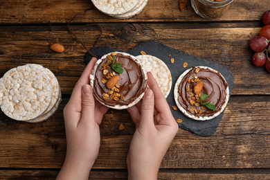 Photo of Woman holding puffed rice cake with chocolate spread at wooden table, top view