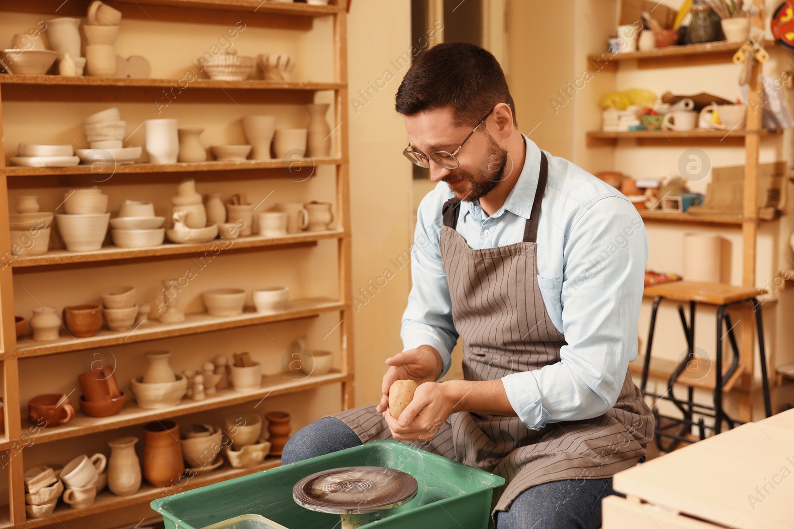 Photo of Man crafting with clay indoors. Different ceramics and potter's wheel in workshop