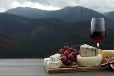 Photo of Different types of delicious cheeses, fruits and wine on wooden table against mountain landscape. Space for text