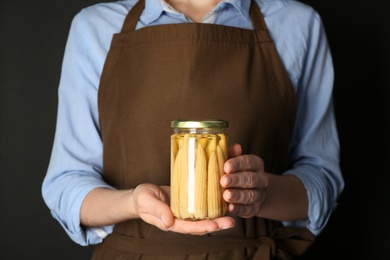Woman holding glass jar of pickled baby corn on black background, closeup