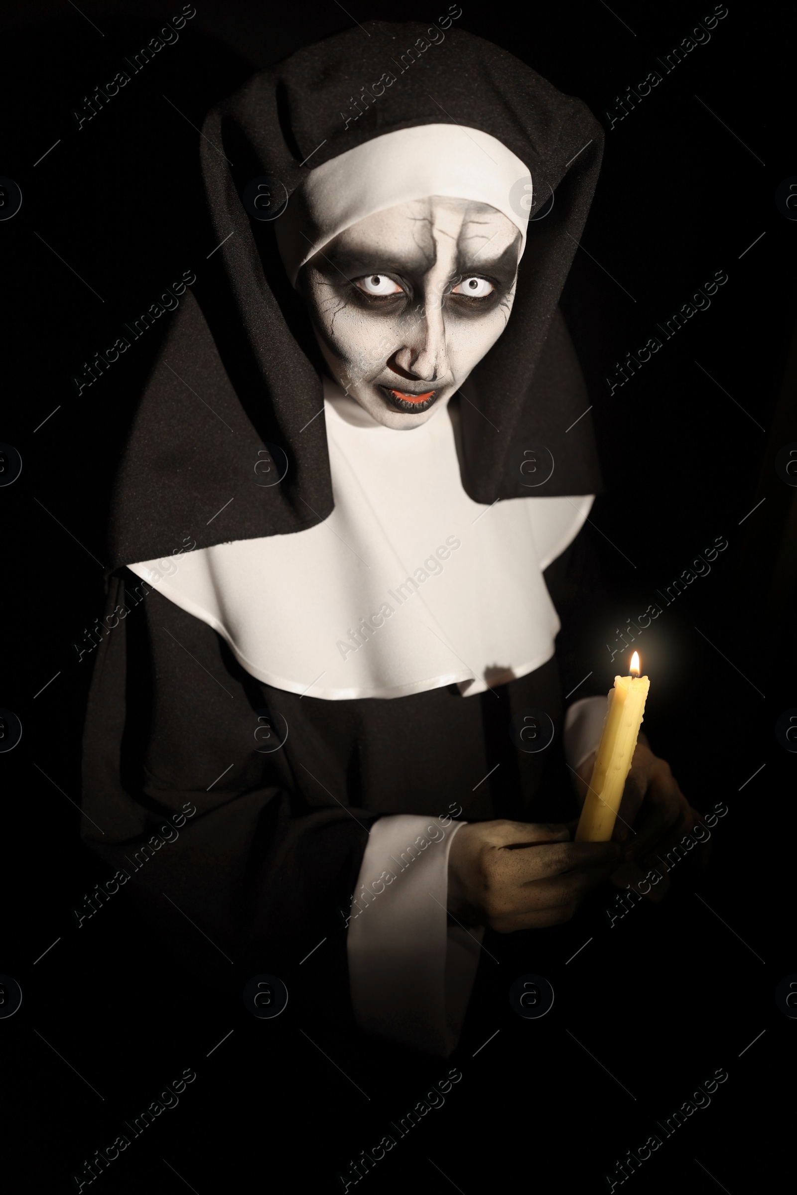 Photo of Scary devilish nun with burning candle on black background. Halloween party look