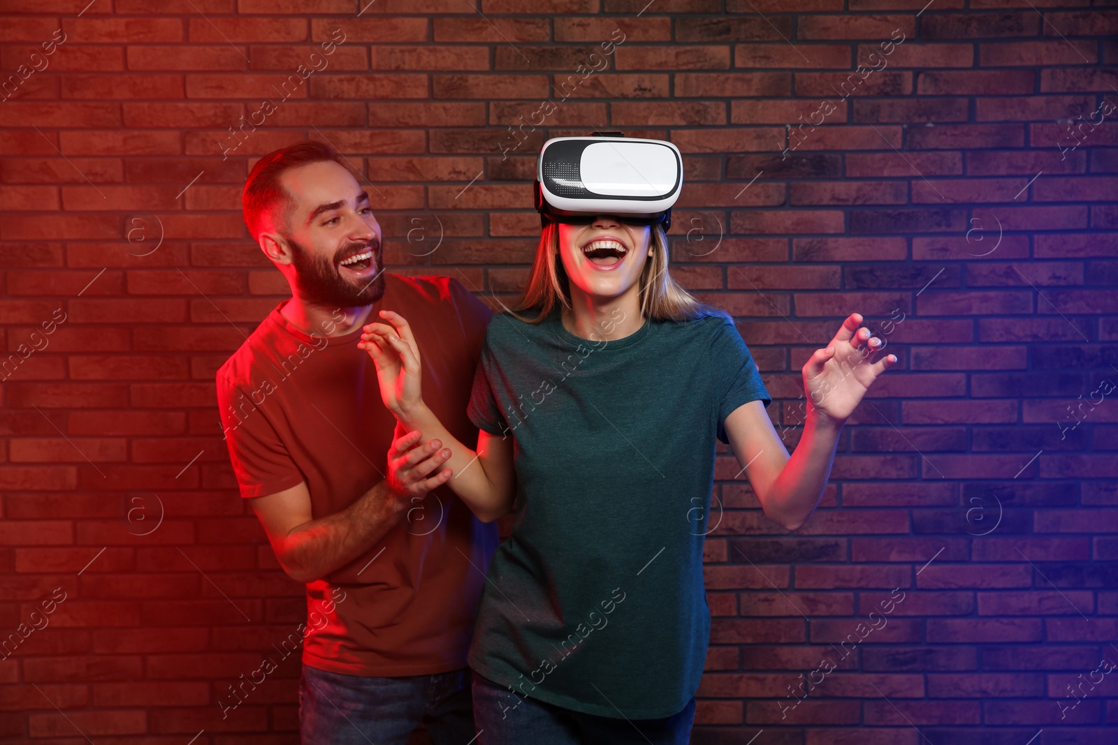 Photo of Emotional woman playing video games with VR headset and happy man near brick wall