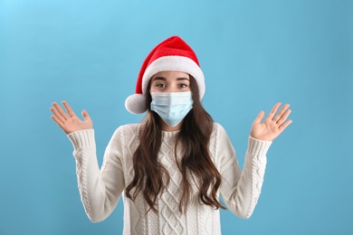 Photo of Beautiful emotional woman wearing Santa Claus hat and medical mask on light blue background