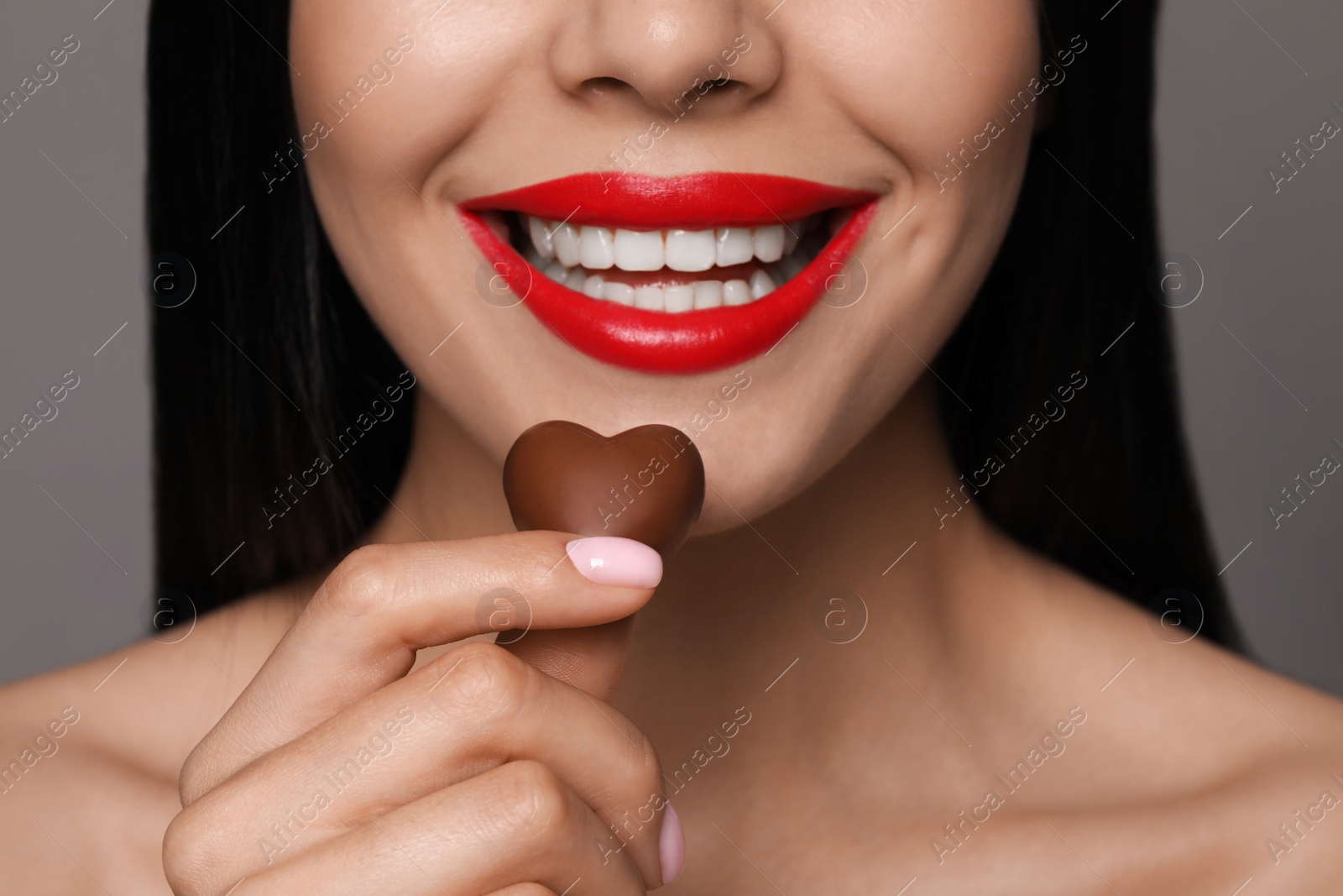 Photo of Young woman with red lips holding heart shaped chocolate candy on grey background, closeup