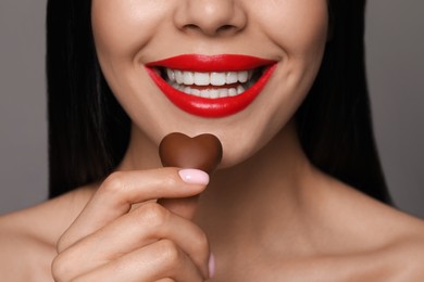 Young woman with red lips holding heart shaped chocolate candy on grey background, closeup