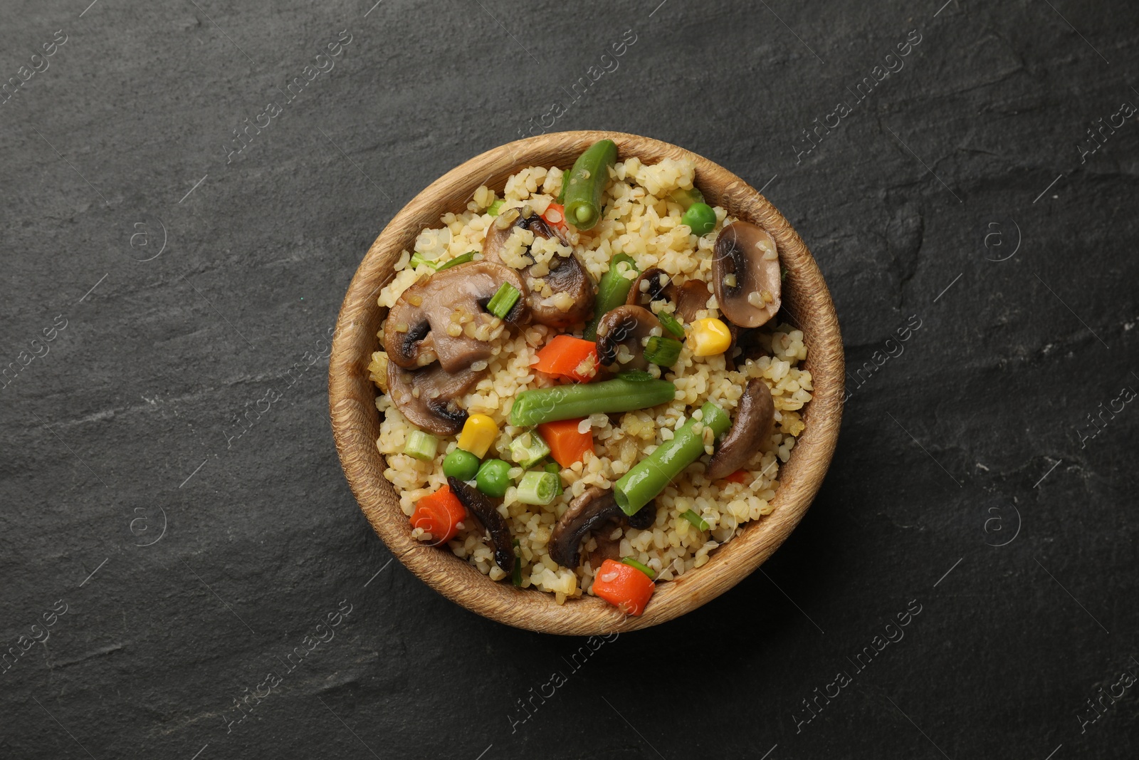 Photo of Delicious bulgur with vegetables and mushrooms in bowl on black table, top view