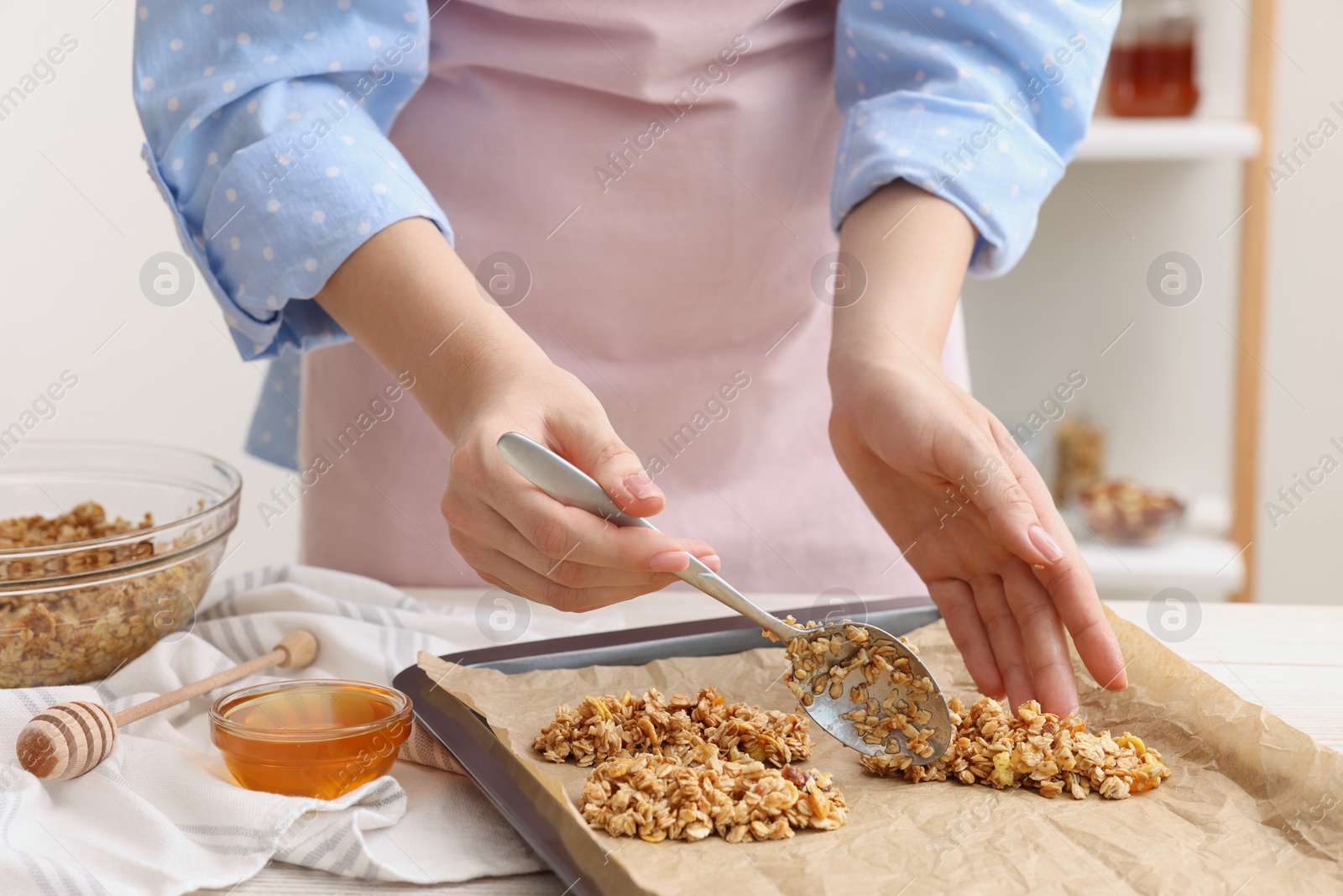 Photo of Making granola bars. Woman putting mixture of oat flakes, dry fruits and other ingredients onto baking tray at table in kitchen, closeup