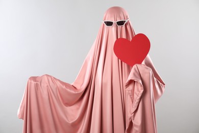 Photo of Cute ghost. Woman in pink sheet with sunglasses and red heart on light grey background