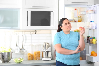 Overweight woman with fresh apple near open refrigerator in kitchen, space for text. Healthy diet