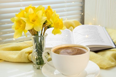 Photo of Beautiful yellow daffodils in vase, cup of coffee, book and festive lights on windowsill