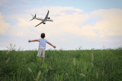 Boy in green field looking at airplane flying in sky, back view