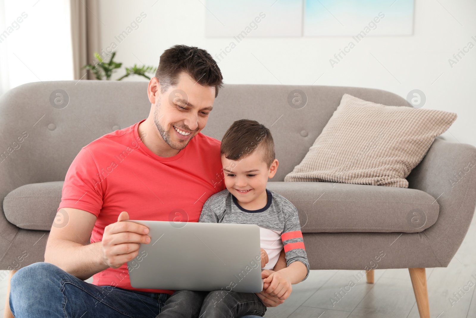 Photo of Boy and his father with laptop sitting near the sofa on floor. Family time