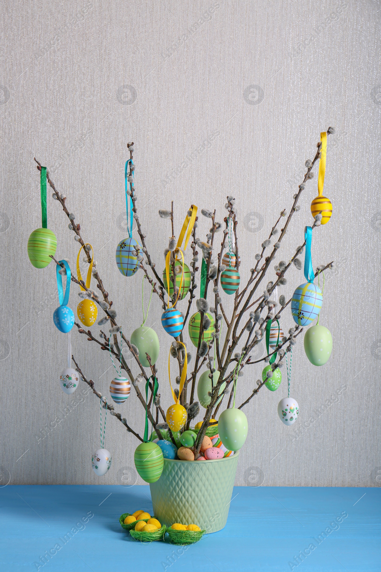 Photo of Beautiful willow branches with painted eggs in pot on light blue table. Easter decor