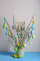 Photo of Beautiful willow branches with painted eggs in pot on light blue table. Easter decor